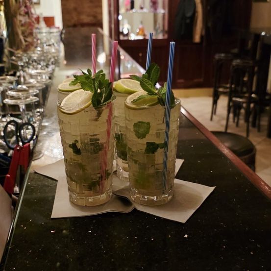 Drei Cocktails mit Strohhalmen und Garnierung auf einer Bar. Gläser gefüllt mit Eis, Minze und Zitronenscheiben.