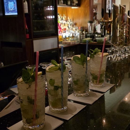 Eine Bartheke mit vier Mojitos darauf. Jeder Drink ist mit Minzblättern und Limettenscheiben garniert. Hinter der Theke befindet sich eine Ausstellung von Spirituosenflaschen und anderen Barutensilien.