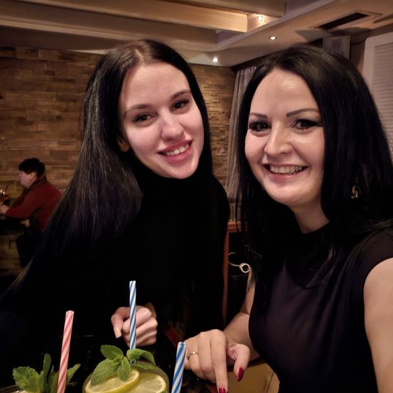 Zwei Frauen lächeln in die Kamera. Sie halten Getränke mit Minzblättern und Zitronenscheiben. Sie sind in einem Restaurant mit einer Ziegelsteinwand und einer Person, die im Hintergrund sitzt.