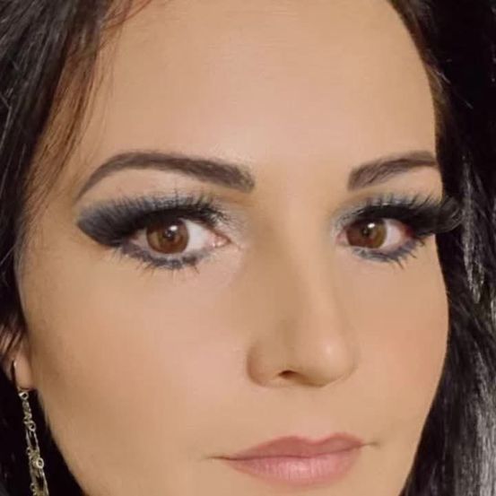 Ein Nahaufnahme des Gesichts einer Frau mit dunklem Haar und braunen Augen. Sie trägt Make-up, einschließlich Eyeliner, Wimperntusche und Lippenstift. Sie trägt ein schwarzes Oberteil und Perlenohrringe.