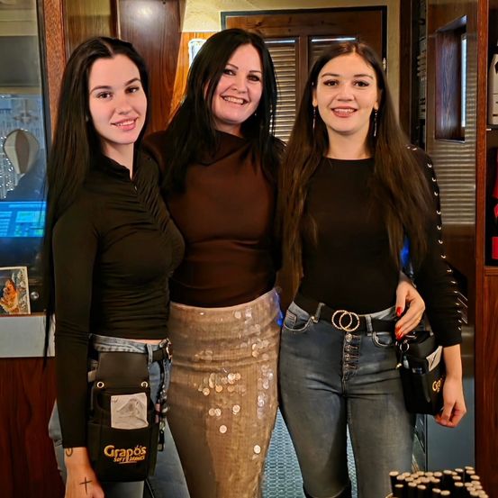 Drei Frauen posieren drinnen zusammen. Eine trägt ein schwarzes Shirt, eine schwarze Tasche und Jeans. Eine andere trägt ein braunes Shirt und eine Paillettenrock. Die dritte trägt ein schwarzes Shirt und hält eine schwarze Tasche. Sie lächeln in die Kamera.