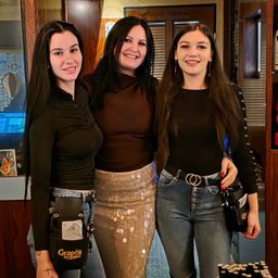 Drei Frauen posieren drinnen zusammen. Eine trägt ein schwarzes Shirt, eine schwarze Tasche und Jeans. Eine andere trägt ein braunes Shirt und eine Paillettenrock. Die dritte trägt ein schwarzes Shirt und hält eine schwarze Tasche. Sie lächeln in die Kamera.