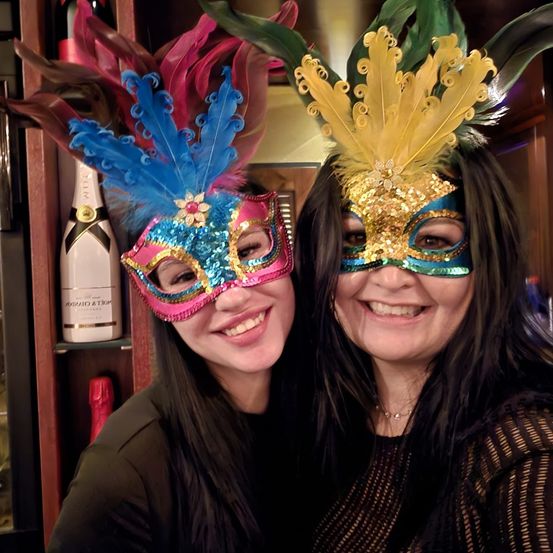 Zwei Frauen in farbenfrohen Masken stehen eng beieinander und lächeln für ein Foto. Dahinter befindet sich ein Regal mit einer Flasche Champagner und einer weiteren Flasche.