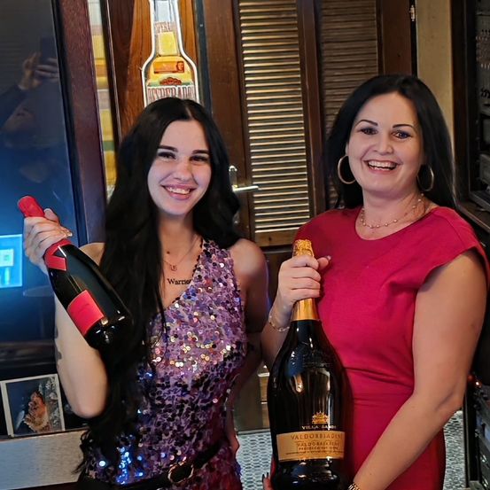 Zwei Frauen stehen nebeneinander, lächeln und halten Champagnerflaschen. Eine trägt ein Paillettenkleid, die andere ein rotes Kleid. Hinter ihnen befindet sich eine Tür mit einem Fenster, das eine Person und eine Tequilaflasche reflektiert.