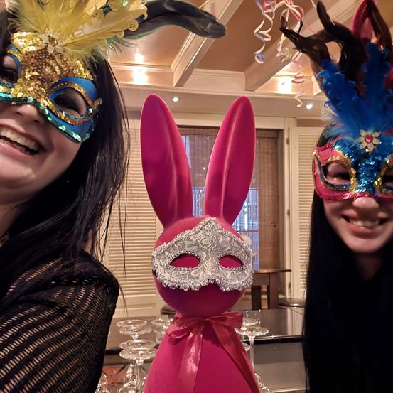 Zwei Frauen in Kostümen posieren mit einem großen rosa Hasen, der eine silberne Maske trägt. Sie tragen bunte Masken mit Federn. Hinter ihnen ist ein Tisch mit Weingläsern zu sehen.