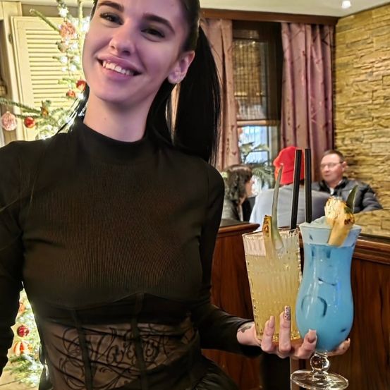 Eine Frau in einem schwarzen Kleid hält zwei Cocktails vor einem Weihnachtsbaum und einem Mann mit rotem Hut und Brille.