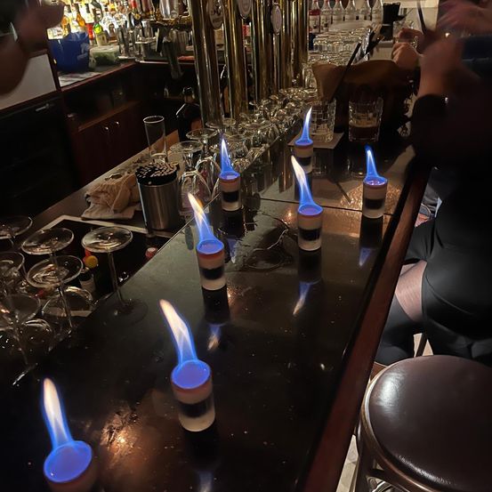 Eine Bartheke mit sechs Getränken in Flammen, blaue Flammen auf der Oberseite, umgeben von Gläsern und Flaschen, und eine Person sitzt in der Nähe.