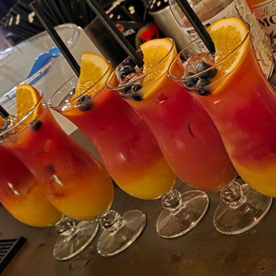 Vier Gläser mit einem geschichteten Cocktail, verziert mit Orangenscheiben und Blaubeeren, stehen auf einem Tisch mit unscharfem Hintergrund.