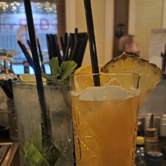 Zwei Getränke stehen auf einer hölzernen Bar, garniert mit Minze, Zitronenscheiben und Ananas. Die Getränke haben schwarze Strohhalme und ein Glas mit strukturiertem Design. Im Hintergrund ist eine verschwommene Ansicht eines Fensters und einer Person zu sehen.