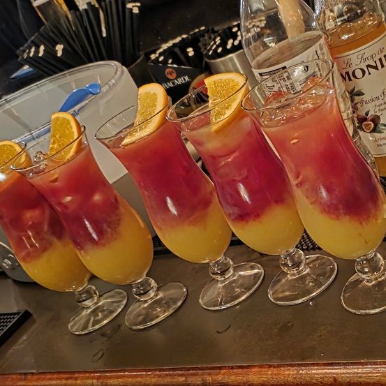 Fünf Gläser mit rosa und gelben Cocktailgetränken mit Eis und Orangenscheiben stehen auf einer Bar. Dahinter befinden sich Flaschen mit Spirituosen und ein Behälter.