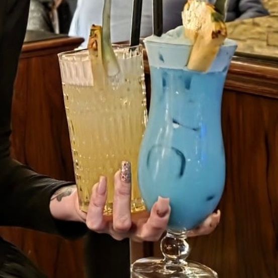 Eine Person hält zwei Getränke in einer Bar, eines gelb und eines blau, beide mit Ananas garniert. Im Hintergrund sitzen zwei Personen an einem Tisch.