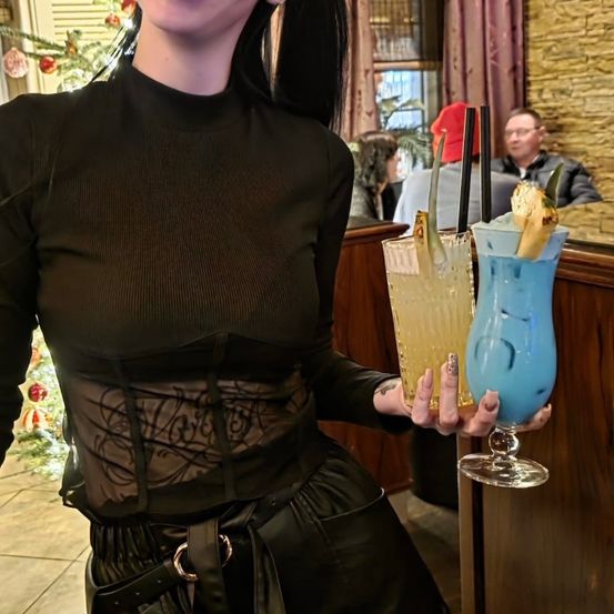 Eine Frau in einem schwarzen Oberteil und Lederrock hält zwei Drinks mit Ananas vor einer Theke. Dahinter ist ein Mann in einem roten Hut und einer Brille zu sehen, zusammen mit einem Weihnachtsbaum.