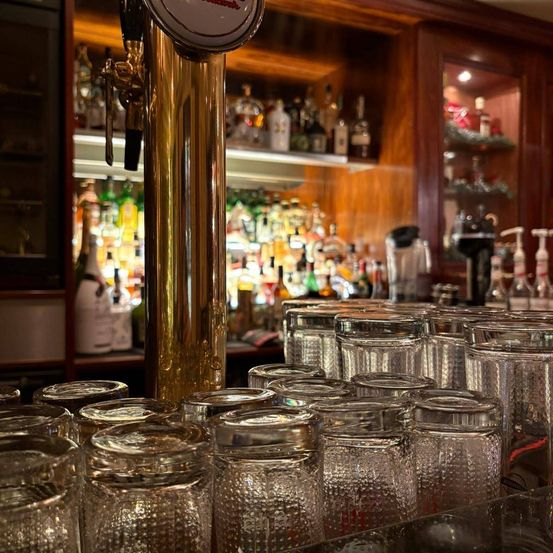 Eine Bar mit einem Messinghahn und einem Regal mit vielen Alkoholflaschen. Gläserne Gläser und Tassen sind auf dem Tresen angeordnet.