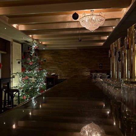 Ein Barbereich mit einem langen, glänzenden Tresen und einem festlich geschmückten Weihnachtsbaum im Hintergrund. Die Decke ist mit einer Kristallleuchte und dekorativen Elementen geschmückt.