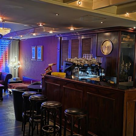 Ein moderner Bar-Tresen ist mit einer Kaffeemaschine, Gläsern und einer Uhr geschmückt. Bilderrahmen sind an einer lila Wand montiert. Eine Kronleuchter hängt von der Decke.