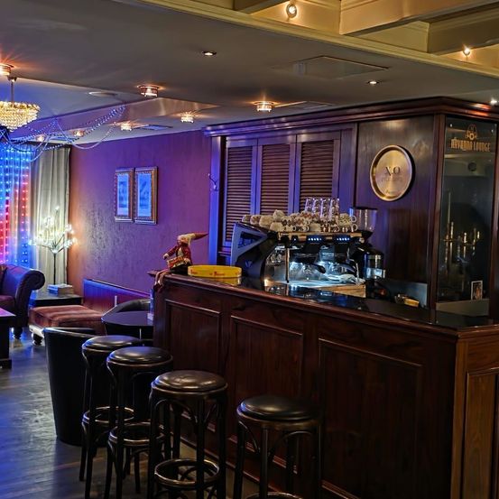 Ein moderner Bar-Tresen ist mit einer Kaffeemaschine, Gläsern und einer Uhr geschmückt. Bilderrahmen sind an einer lila Wand montiert. Eine Kronleuchter hängt von der Decke.