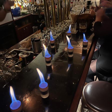 Eine Bartheke mit brennenden Kerzen in Shotgläsern. Es gibt Weingläser, Flaschen und andere Gegenstände. Eine Person sitzt und hält eine Zigarette.