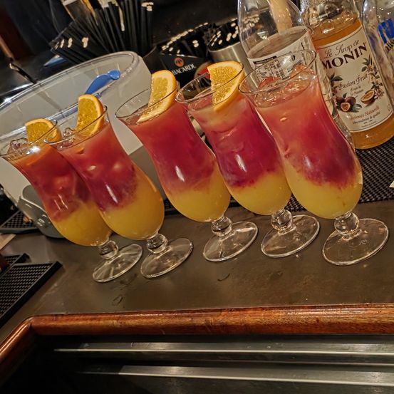 Fünf hohe Gläser mit einem rosa und orangefarbenen geschichteten Getränk, das mit Zitronenscheiben garniert ist, sind auf einem Tresen angeordnet. Im Hintergrund sind Flaschen und Utensilien zu sehen.