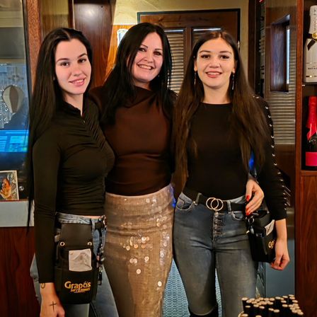 Drei Frauen stehen lächelnd zusammen in einem Raum. Eine trägt ein braunes Oberteil und eine Hose mit einem Paillettenrock, eine andere ein schwarzes Langarmshirt mit einer Tasche und die dritte Jeans und ein schwarzes Shirt. Hinter ihnen steht ein Holzschrank mit einer Flasche Wein.