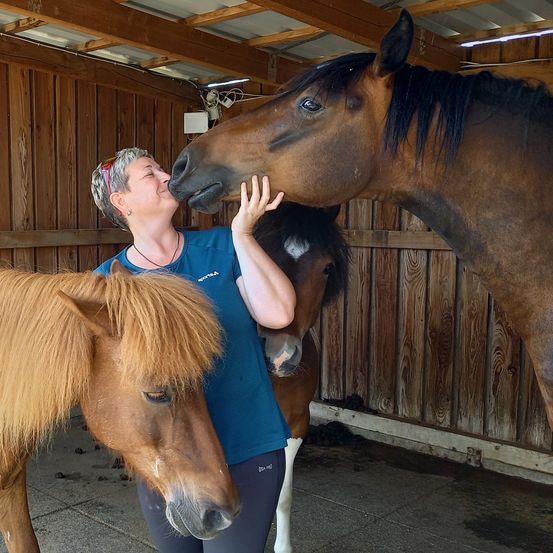Eine Frau steht in einem Stall mit zwei Pferden, von denen eines sie küsst.