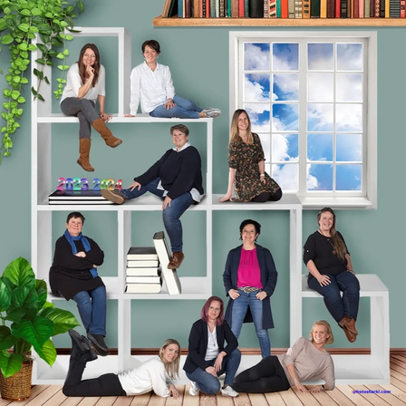 Eine Gruppe von Frauen posiert in einer thematischen Umgebung mit einem Regal, Büchern und einem Fenster, das einen Himmel zeigt. Sie sitzen oder liegen auf Regalen und dem Boden.