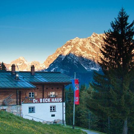 Ein traditionelles Holzhaus mit Dr. Beck Haus darauf geschrieben. Das Haus ist von hohen Bäumen umgeben und im Hintergrund befinden sich Berge.