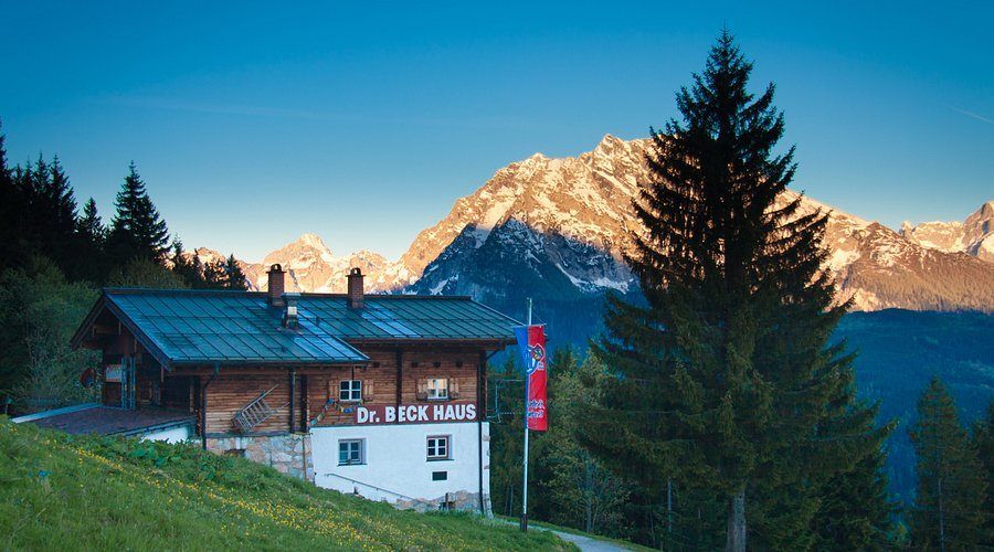 Ein traditionelles Holzhaus mit Dr. Beck Haus darauf geschrieben. Das Haus ist von hohen Bäumen umgeben und im Hintergrund befinden sich Berge.
