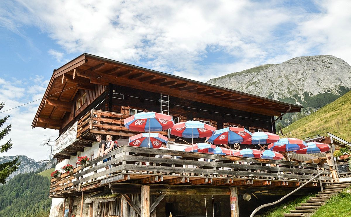 Eine Berghütte mit einer Sitzgelegenheit im Freien unter mehreren roten und blauen Sonnenschirmen, mit einem Berg im Hintergrund.