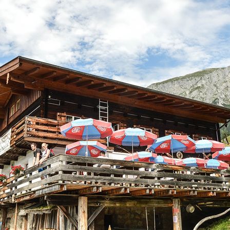 Eine Berghütte mit einer Sitzgelegenheit im Freien unter mehreren roten und blauen Sonnenschirmen, mit einem Berg im Hintergrund.