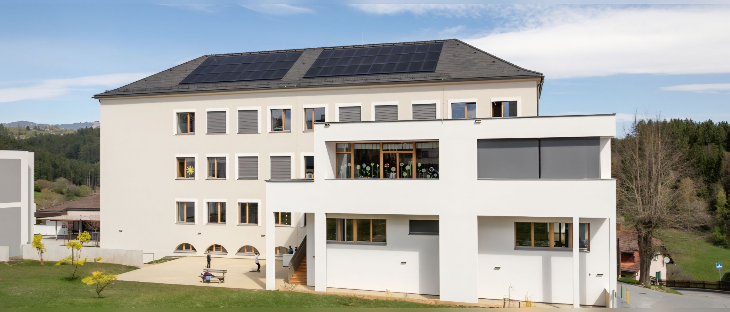 Ein weißes Gebäude mit Solarmodulen auf dem Dach und Glasfenstern. Eine Person läuft auf dem Rasen vor dem Gebäude.