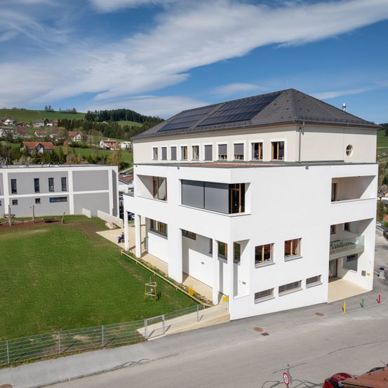 Ein großes weißes Gebäude mit flachem Dach und Solarpanelen. Das Gebäude ist von einem Rasenfeld und einer Straße umgeben. Ein kleineres Gebäude steht links.