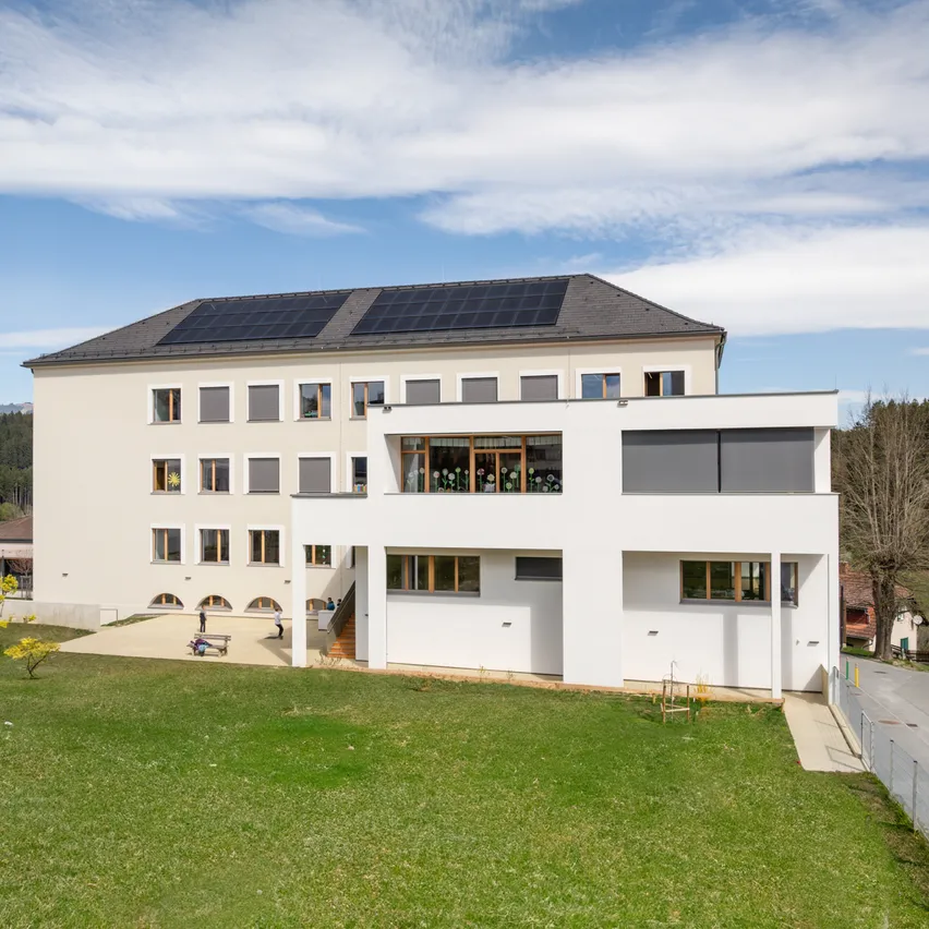 Ein weißes Gebäude mit Solarpanelen auf dem Dach und Fenstern mit Rollläden. Einige Personen sind draußen.