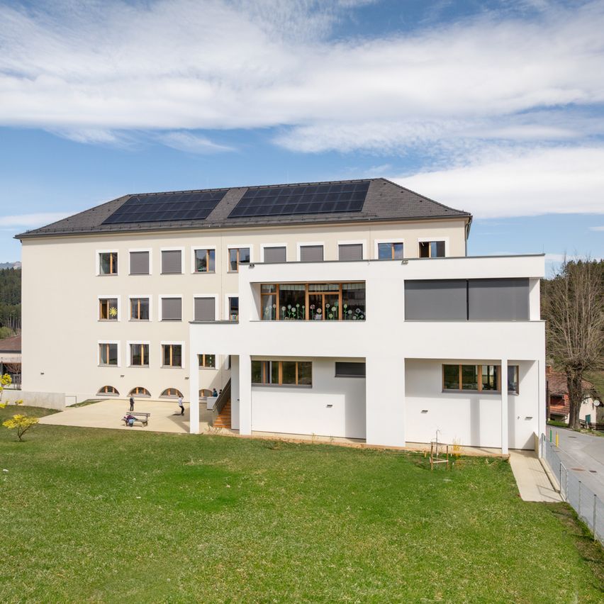 Ein weißes Gebäude mit Solarpanelen auf dem Dach und Fenstern mit Rollläden. Einige Personen sind draußen.