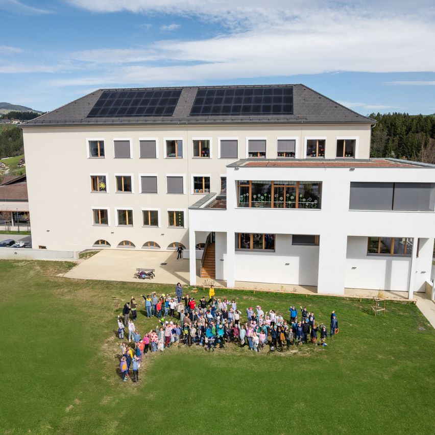 Luftaufnahme eines großen, modernen Gebäudes mit Solarpanelen auf dem Dach, umgeben von einer großen Gruppe von Menschen auf einem grünen Feld.
