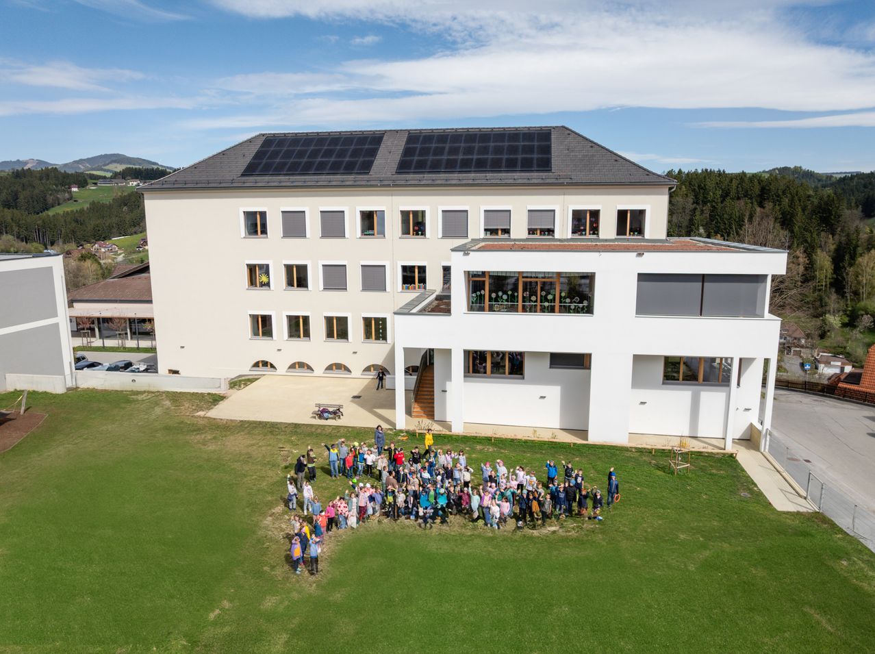 Luftaufnahme eines großen, modernen Gebäudes mit Solarpanelen auf dem Dach, umgeben von einer großen Gruppe von Menschen auf einem grünen Feld.