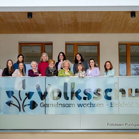 Eine Gruppe von Frauen in Winterkleidung posiert auf einem Balkon vor einem Schild mit der Aufschrift 'Volksschule Geminsam wachsen'.