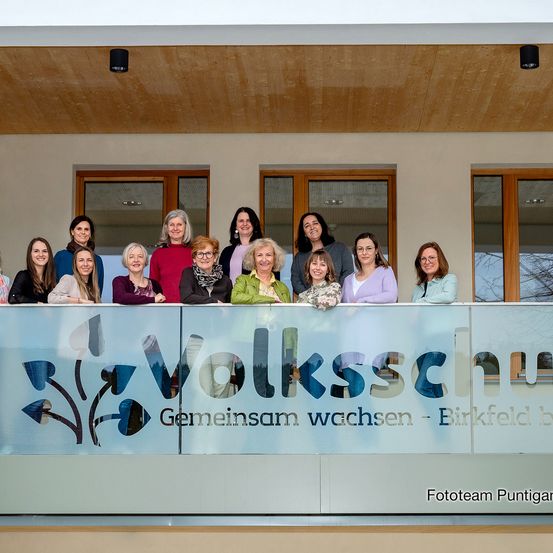 Eine Gruppe von Frauen in Winterkleidung posiert auf einem Balkon vor einem Schild mit der Aufschrift 'Volksschule Geminsam wachsen'.