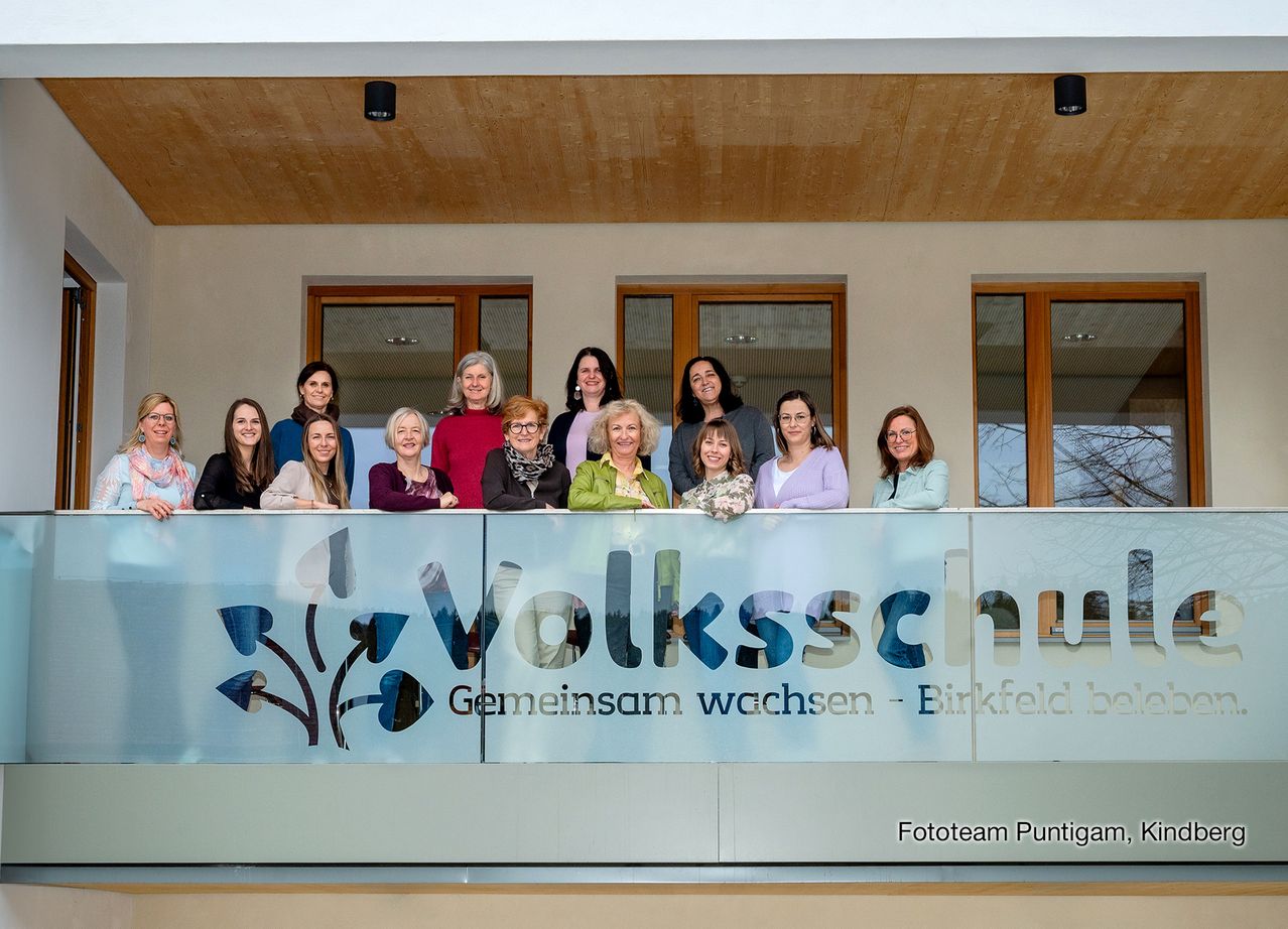Eine Gruppe von Frauen in Winterkleidung posiert auf einem Balkon vor einem Schild mit der Aufschrift 'Volksschule Geminsam wachsen'.