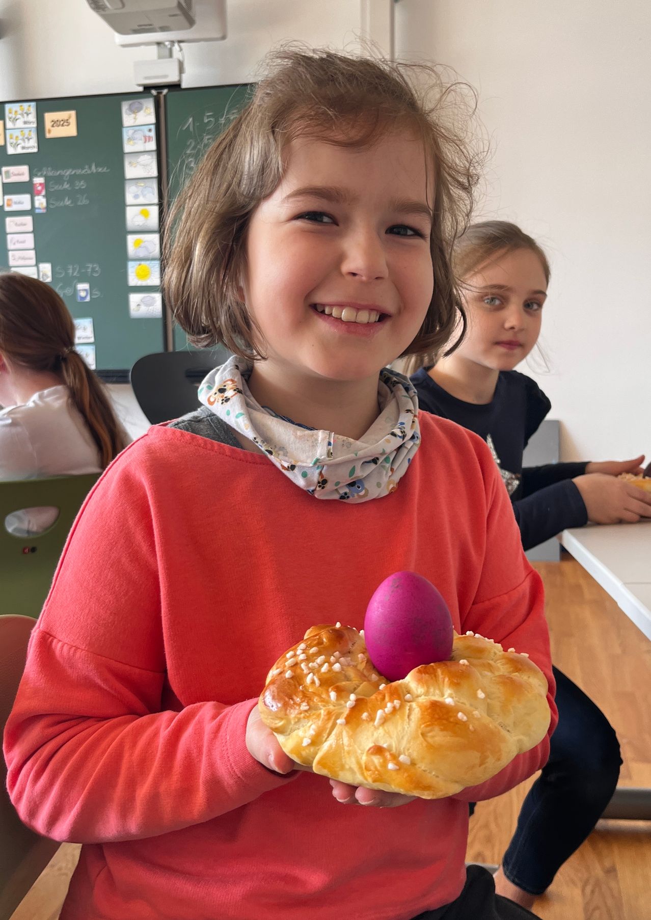 Ein Mädchen in einem roten Shirt hält ein rosa Osterei auf einem süßen Brot in einem Klassenzimmer.