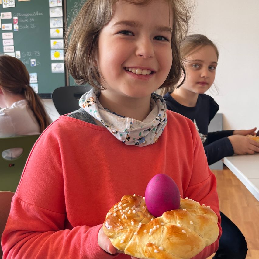 Ein Mädchen in einem roten Shirt hält ein rosa Osterei auf einem süßen Brot in einem Klassenzimmer.