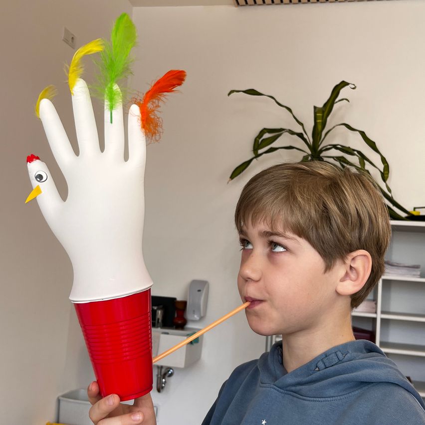 Ein Junge mit einer blauen Kapuzenjacke hält eine Handpuppe in den Händen, die er an seinen Mund hält und hineinbläst.