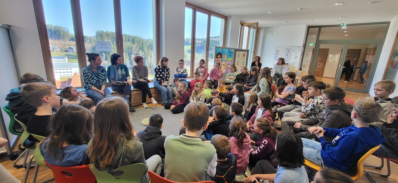 Eine Gruppe von Kindern und Erwachsenen versammelt sich in einem Raum mit großen Fenstern, die auf Bäume und Berge draußen schauen.