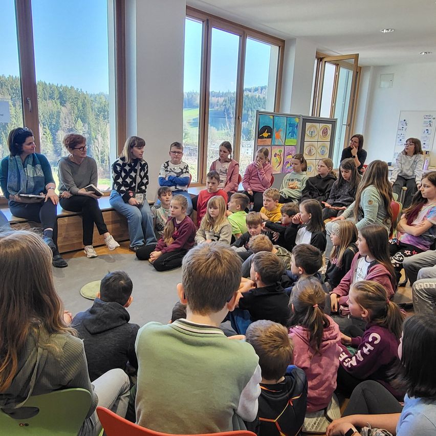 Eine Gruppe von Kindern und Erwachsenen versammelt sich in einem Raum mit großen Fenstern, die auf Bäume und Berge draußen schauen.