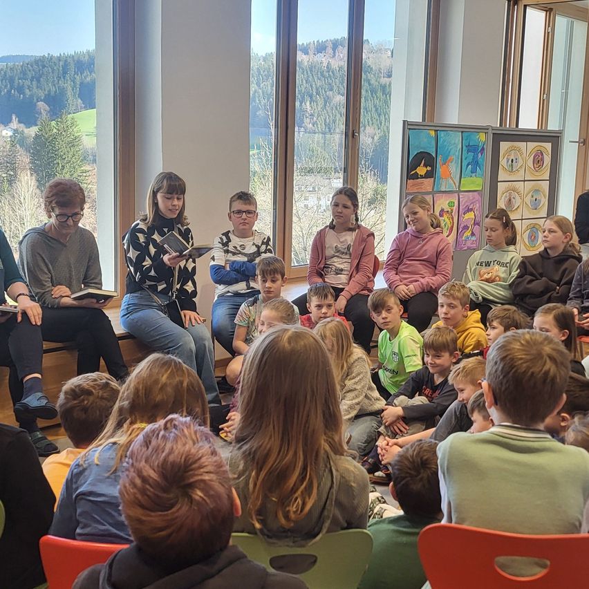 In einem Klassenzimmer mit großen Fenstern versammeln sich eine Gruppe von Kindern und Erwachsenen. Sie sitzen im Kreis auf dem Boden. Einige Kinder halten Bücher in den Händen, andere schauen zu. Eine Frau mit Brille steht in der Mitte, hält ein Buch und liest laut vor.