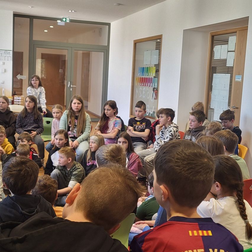 Eine Gruppe von Schülern sitzt im Kreis, wahrscheinlich in einem Klassenzimmer. Einige tragen eine Brille und andere ein Armband. Eine Frau im Hintergrund steht, wahrscheinlich als Lehrerin. Eine Glastür befindet sich auf der linken Seite und eine Tafel mit angehefteten Papieren auf der rechten.
