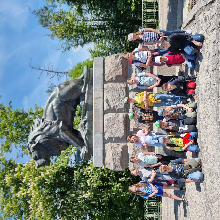 Eine Gruppe von Kindern posiert für ein Foto vor einer Löwenstatue an einem sonnigen Tag. Bäume und ein Zaun sind im Hintergrund zu sehen.