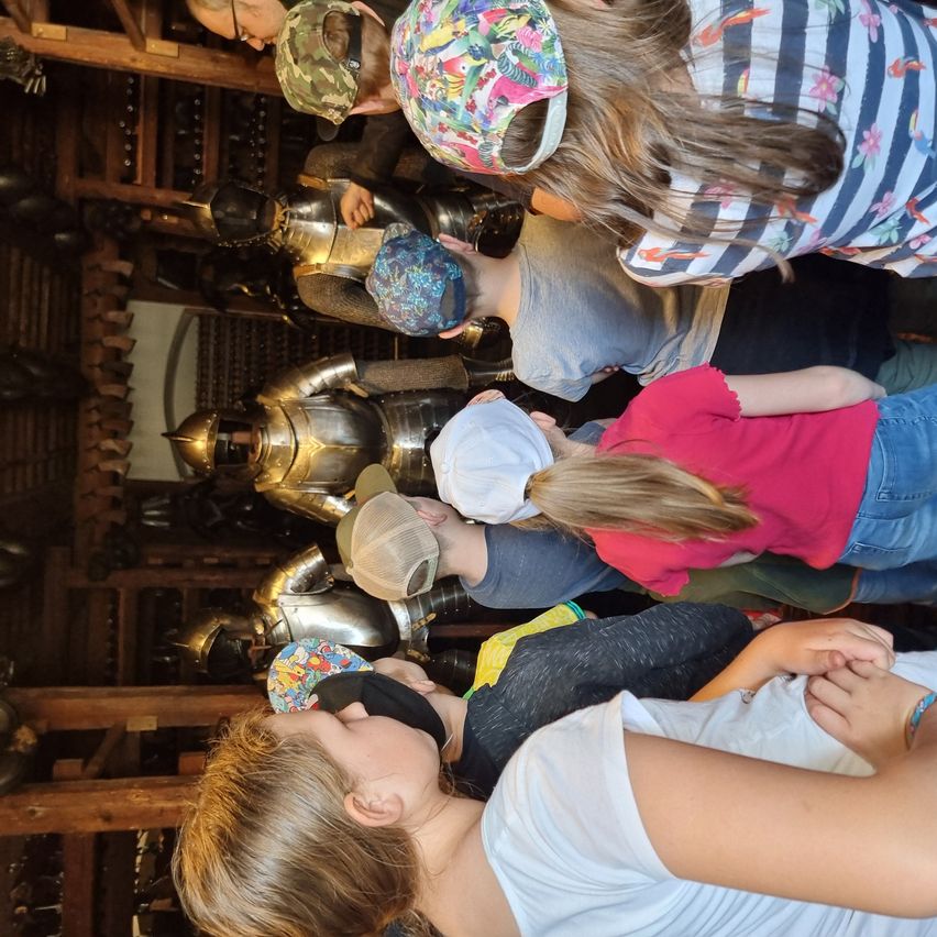 Eine Gruppe von Menschen, einige mit Mützen, versammelt sich um eine Ausstellung von Ritterrüstungen und betrachtet sie aufmerksam. Sie befinden sich in einem holzstrukturierten Raum.