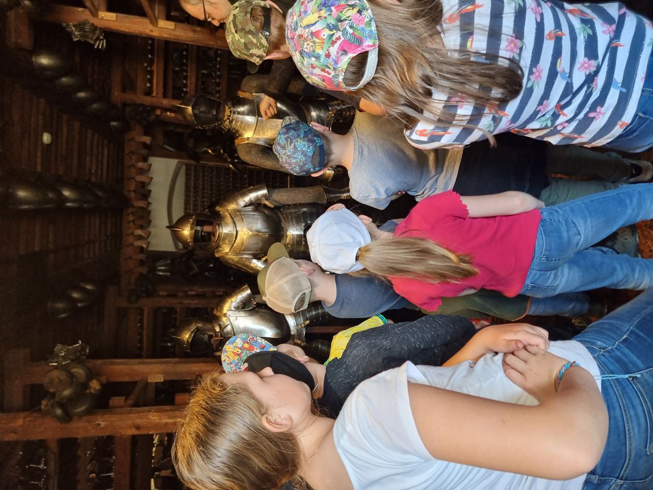 Eine Gruppe von Menschen, einige mit Mützen, versammelt sich um eine Ausstellung von Ritterrüstungen und betrachtet sie aufmerksam. Sie befinden sich in einem holzstrukturierten Raum.