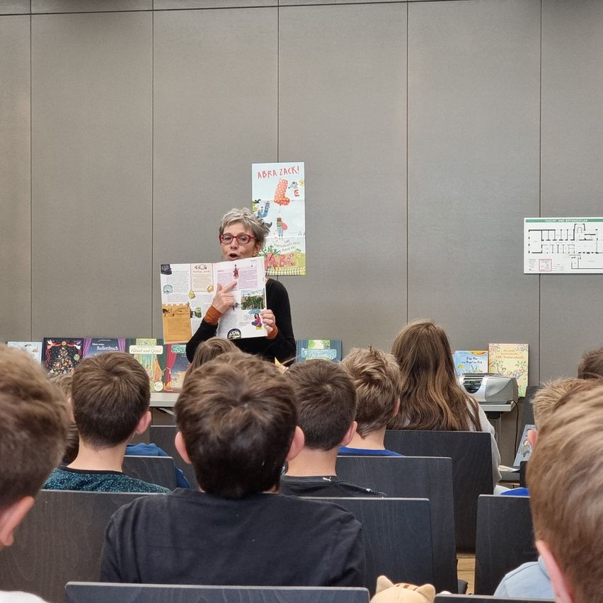 Eine Frau liest ein Buch für eine Klasse von Kindern vor, die aufmerksam zuhören. Hinter ihr steht ein Poster mit der Aufschrift 'Abra Zack!'.