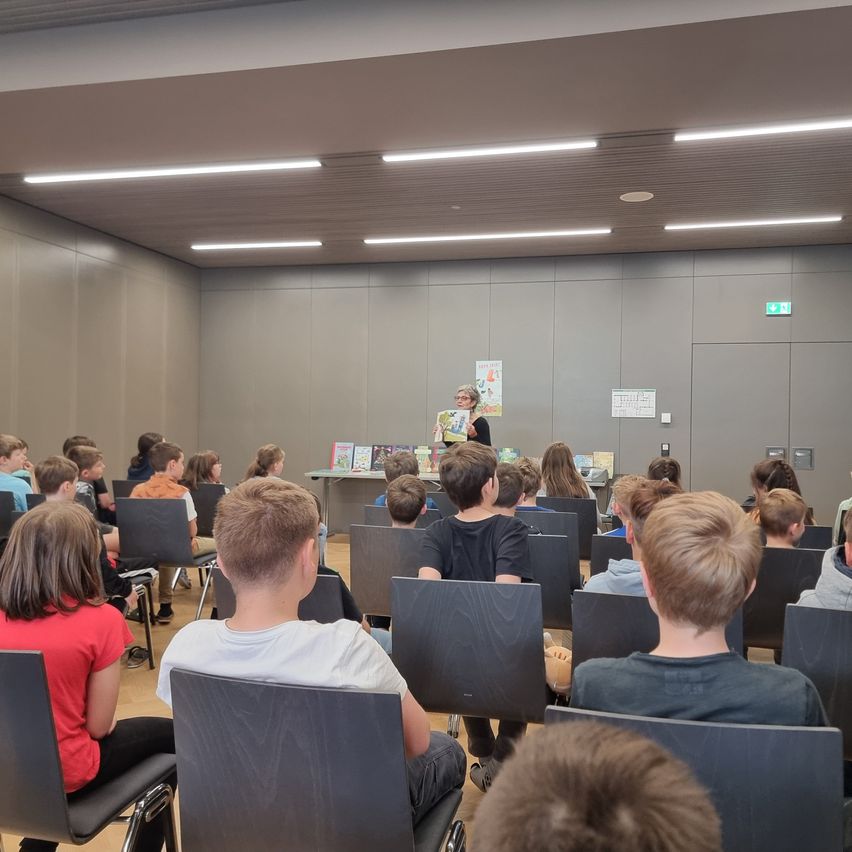 Eine Frau steht an einem Podium in einem Klassenzimmer, umgeben von Kindern, die auf Stühlen sitzen und zuhören. Das Klassenzimmer hat graue Wände und Deckenleuchten.